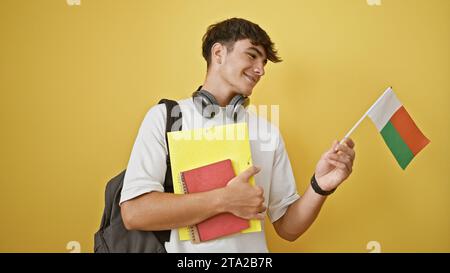 Portrait captivant d'un jeune étudiant hispanique adolescent, arborant fièrement le drapeau de madagascar et des livres sur un fond jaune vif isolé Banque D'Images