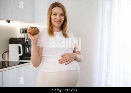 Une femme enceinte aux cheveux blonds souriants tient une pomme et met sa main sur son ventre dans la cuisine. Régime alimentaire pendant la grossesse. Banque D'Images