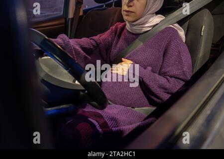 Femme arabe musulmane enceinte conduisant la voiture la nuit portant des vêtements d'hiver, hijab et ceinture de sécurité conduisant en toute confiance et se sentant en sécurité Banque D'Images