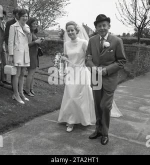 Années 1960, historique, mariage, un père fier, regardant dapper dans un costume avec fleur d'œillet et chapeau, accompagne sa fille, la mariée, portant un fer à cheval chanceux, en haut du chemin, regardé par des invités enthousiastes, Angleterre, Royaume-Uni. Banque D'Images