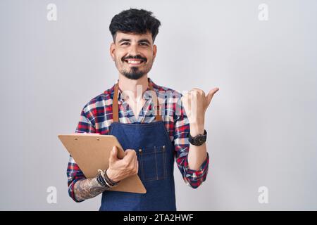 Jeune homme hispanique avec barbe portant un tablier de serveur tenant le presse-papiers pointant vers l'arrière derrière avec la main et les pouces vers le haut, souriant confiant Banque D'Images