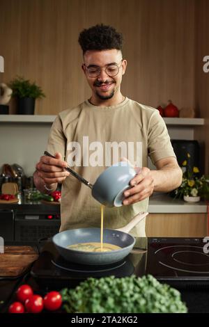 Plan vertical moyen d'un jeune chef souriant du Moyen-Orient versant des œufs battus sur une poêle tout en posant près de la cuisinière Banque D'Images