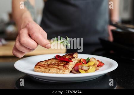 Gros plan de la main d'un homme méconnaissable mettant du vert sur le dessus d'un steak fraîchement cuit avec des légumes autour, espace de copie Banque D'Images