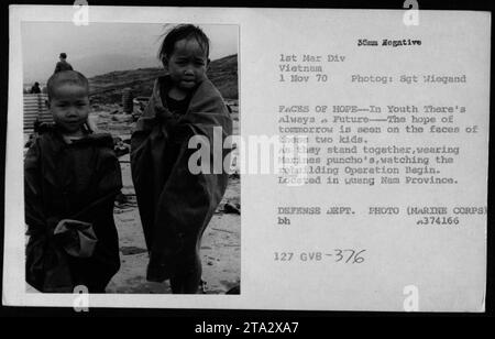 Deux enfants vietnamiens, portant les ponchos de Marine, observent l'opération de reconstruction dans la province de Luang Nam. Leur jeunesse et leurs expressions d'espoir symbolisent l'avenir au milieu de la tourmente de la guerre du Vietnam. Photographié par le sergent Wiegand le 1 novembre 1970. Photo du ministère de la Défense (corps des Marines). Banque D'Images