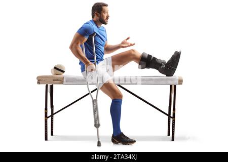 Joueur de football avec une béquille et un pied blessé assis sur un lit de physiothérapie isolé sur fond blanc Banque D'Images