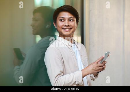 Jeune homme asiatique avec des écouteurs sans fil tenant un téléphone intelligent Banque D'Images