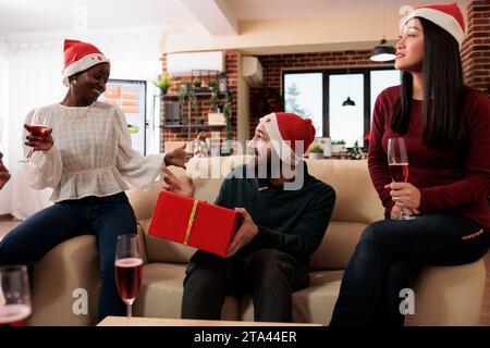 Homme excité avec boîte cadeau enveloppée festive devinant le père noël secret tout en célébrant noël dans le bureau. Collègues joyeux dans des chapeaux de Noël échangeant des cadeaux et profitant des vacances du nouvel an Banque D'Images
