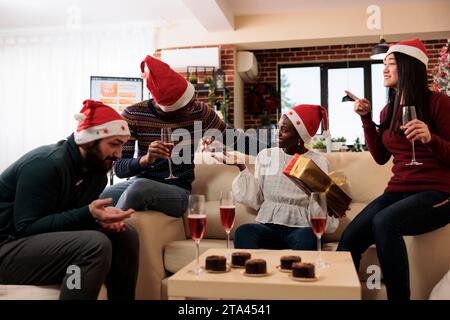 Joyeux employé afro-américain excité tenant le cadeau de Noël et devinant le père noël secret dans le bureau. Collègues divers buvant de l'alcool et partageant des cadeaux de noël Banque D'Images