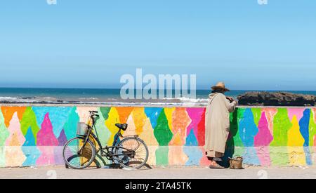 Essaouira, Maroc - 17 septembre 2022 : Homme marocain en habit traditionnel avec son vélo appuyé sur un mur coloré au bord de la côte d'Essaouira Banque D'Images