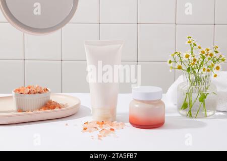 Vue de face d'un tube blanc et pot sans étiquette disposés avec un bol de sel rose de l'himalaya et pot de fleurs. Le sel rose de l'Himalaya est un type de sel tha Banque D'Images