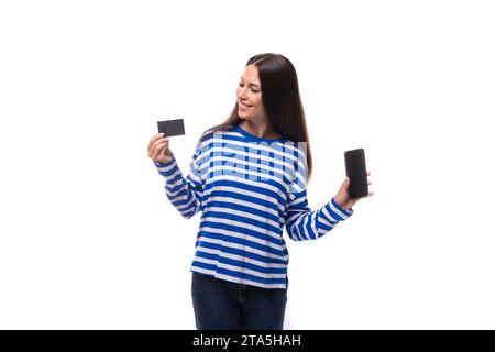 jeune femme brune européenne confiante vêtue de vêtements bleus rayés annonce carte bancaire et téléphone intelligent Banque D'Images