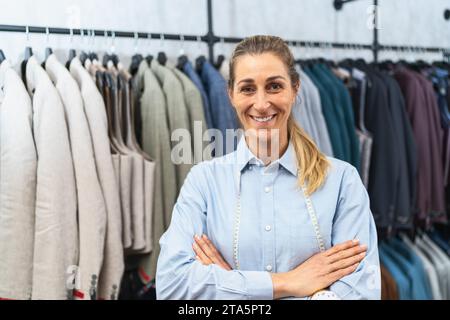 Tailleur confiant avec les bras croisés debout devant les vestes de costume sur des cintres Banque D'Images