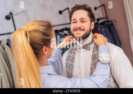 Tailleur ajustant un noeud papillon sur un homme joyeux dans un magasin de mode avec des porte-vêtements Banque D'Images