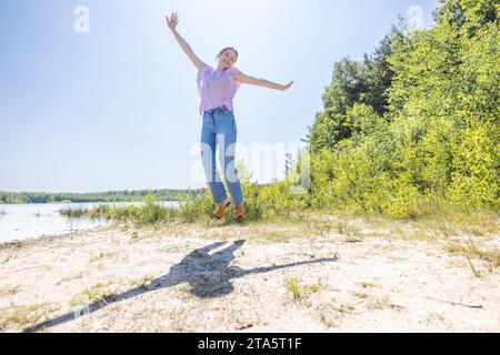 L'image capture un moment exubérant alors qu'une personne saute joyeusement dans les airs. L'ombre de l'individu est fortement projetée sur le sol, suggérant les conditions lumineuses et claires d'une journée ensoleillée. Ils sont habillés de manière décontractée, suggérant confort et atmosphère détendue. La toile de fond du lac et de la verdure luxuriante contribue à un sentiment de liberté et de connexion avec la nature. Joyful Leap : moment insouciant au bord du lac. Photo de haute qualité Banque D'Images