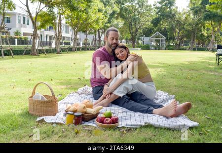 Un jeune couple profite d'un pique-nique romantique par une journée ensoleillée dans un beau parc, assis sur une couverture avec de la nourriture autour d'eux. Banque D'Images