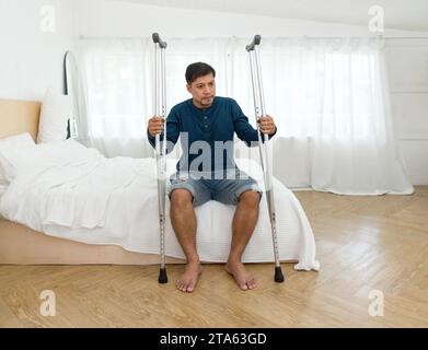 Un homme se repose sur un lit, regardant contemplatif, avec une paire de béquilles penchées à proximité, indiquant une adversité physique récente. Banque D'Images