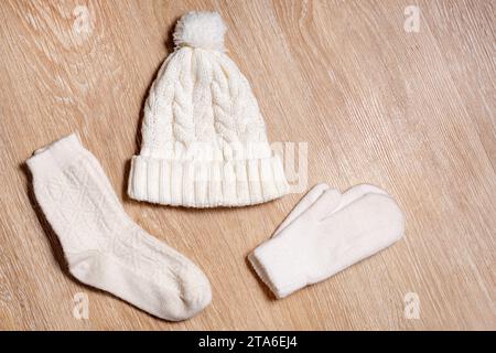 Un bonnet tricoté blanc avec un pompom, des mitaines en laine et des chaussettes reposent sur le fond en bois. Détails des vêtements d'hiver, plat, vue de dessus Banque D'Images