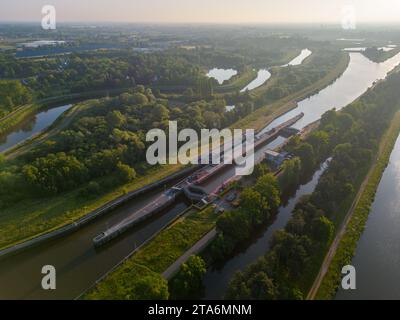 La photo présente une perspective aérienne enchanteresse d'un couloir fluvial en fin de soirée. Le soleil couchant projette une lueur chaude sur la région, illuminant la rivière Snaking, les routes parallèles et la couverture d'arbres verdoyante qui les borde. Comme la voie navigable mène au loin, elle agit comme un guide naturel à travers le paysage luxuriant, tandis que les routes adjacentes parlent de l'intégration de l'infrastructure avec l'environnement. La douce lumière du soir crée de longues ombres et ajoute de la profondeur à cette coexistence pacifique de la nature et de la civilisation. Lumière du soir sur le couloir de la voie navigable. Banque D'Images