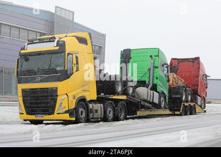 Camion porteur de véhicules Volvo FH jaune transportant deux nouveaux camions Volvo verts et rouges garés dans les chutes de neige hivernales. Salo, Finlande. 24 novembre 2023. Banque D'Images