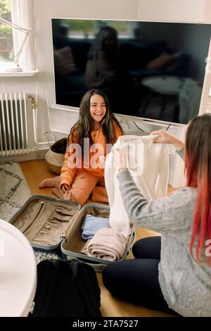 Fille heureuse emballant des vêtements dans le sac avec la mère tout en étant assise dans le salon à la maison Banque D'Images