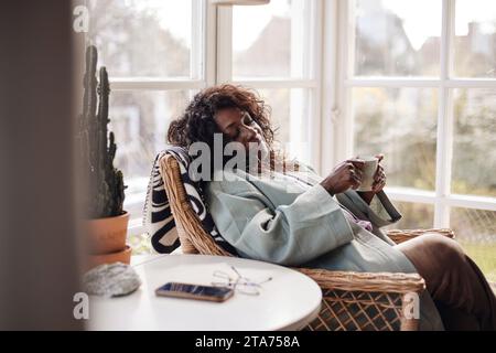 Femme mature tenant une tasse de café tout en se relaxant sur une chaise à la maison Banque D'Images