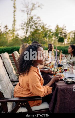 Vue latérale de la femme heureuse riant tout en ayant de la nourriture avec des amis à la table à manger dans la cour arrière Banque D'Images