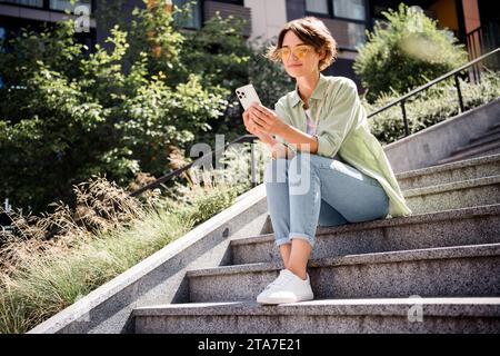 Photo de belle fille mignonne habillée tenue élégante assise sur les escaliers dans le parc lisant télégramme nouvelles instagram post temps libre à l'extérieur Banque D'Images