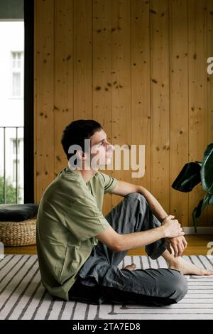 Vue latérale de la jeune femme réfléchie assise sur la moquette à la maison Banque D'Images