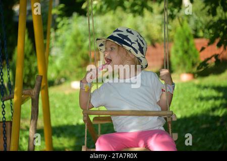 Une petite fille dans un chapeau se balançait sur une balançoire accrochée à un arbre dans le jardin Banque D'Images