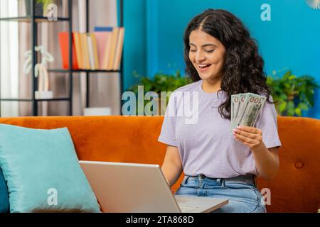 Budget de planification. Riche femme indienne heureuse comptant l'argent comptant utiliser l'ordinateur portable calculer les factures domestiques à la maison. Fille arabe satisfaite des revenus, économise de l'argent pour les vacances planifiées, cadeaux Banque D'Images