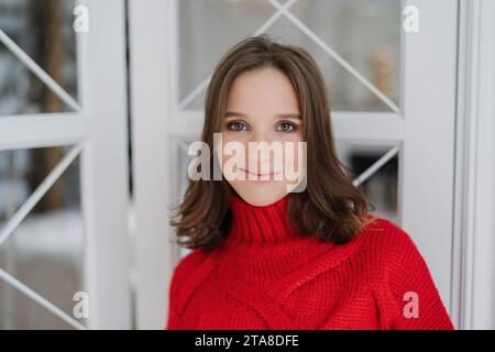 Femme souriante dans un pull à col roulé rouge, à l'air confortable et élégant, avec une fenêtre claire et une scène d'hiver derrière Banque D'Images