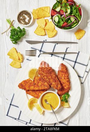 filet de poisson pané cuit au four servi avec de la moutarde jaune sur une assiette blanche, des chips tortilla et une salade fraîche de laitue, tomates, concombre, oignons rouges Banque D'Images