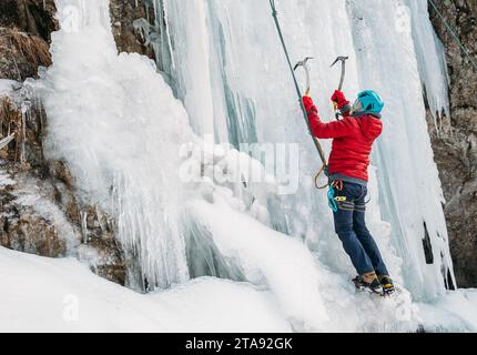 Grimpeur sur glace vêtu de vêtements chauds d'escalade en hiver, d'un harnais de sécurité et d'un casque grimpant une chute d'eau gelée à l'aide de deux haches d'escalade sur glace et de crampons. Agir Banque D'Images