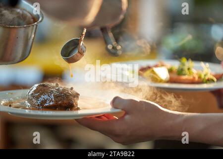 Chef versant la sauce sur le steak. Gros plan sur la cuisine lors d'une Garden party. Deux assiettes à la main, avec steak et saumon, en rétroéclairage coloré, faible profondeur de Banque D'Images