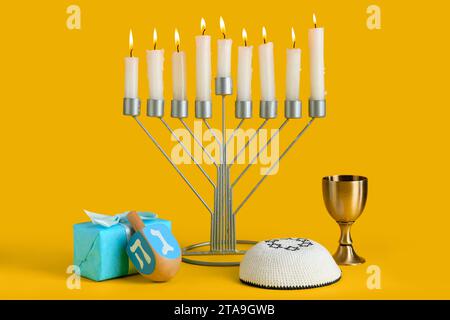 Menorah avec des bougies allumées, boîte-cadeau et symboles Hanukkah sur fond jaune Banque D'Images