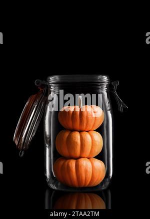 Trois Mini citrouilles décoratives empilées dans un pot de stockage en verre ou de conserve isolé sur noir avec réflexion, avec couvercle ouvert. Banque D'Images