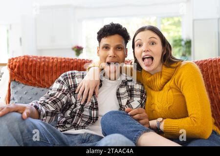 Portrait d'heureux couple diversifié assis sur le canapé, embrassant, faisant des visages drôles à la maison, espace de copie. Expression, loisirs, amour, unité, gesticu Banque D'Images