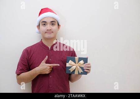 Jeune père Noël asiatique tient et montre une boîte de Noël vert émeraude à la caméra avec l'expression du visage plat. Fond beige isolé. Banque D'Images