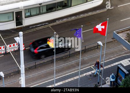 PRODUCTION - 20 novembre 2023, Bade-Württemberg, Weil am Rhein : les drapeaux de l'Allemagne (de gauche à droite), de l'Union européenne et de la Suisse flottent près du poste frontière à Weil am Rhein-Friedlingen tandis qu'une voiture roule derrière eux. La police fédérale intensifie actuellement les contrôles sur les voyageurs, y compris à la frontière avec la Suisse. Le ministre fédéral de l'intérieur, M. Faeser (SPD), a d'abord annoncé le 16 octobre des contrôles stationnaires aux frontières avec la Pologne, la République tchèque et la Suisse pendant dix jours, puis les a prolongés à plusieurs reprises. Elle a justifié cette démarche par la lutte contre l'irregul Banque D'Images