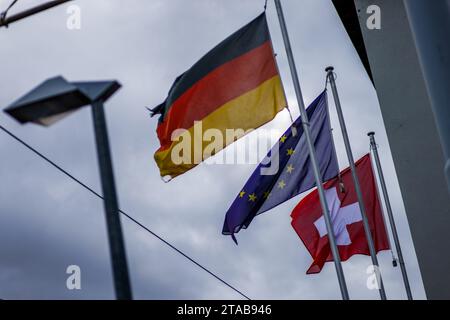 PRODUCTION - 20 novembre 2023, Bade-Württemberg, Weil am Rhein : les drapeaux de l'Allemagne (de gauche à droite), de l'Union européenne et de la Suisse flottent près du poste frontière de Weil am Rhein-Friedlingen. La police fédérale intensifie actuellement les contrôles sur les voyageurs, y compris à la frontière avec la Suisse. Le ministre fédéral de l'intérieur, M. Faeser (SPD), a d'abord annoncé le 16 octobre des contrôles stationnaires aux frontières avec la Pologne, la République tchèque et la Suisse pendant dix jours, puis les a prolongés à plusieurs reprises. Elle a justifié cette démarche par la lutte contre l'immigration clandestine et le trafic de migrants. Banque D'Images