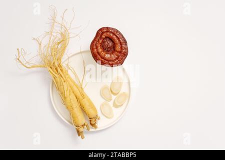 Sur fond blanc, racines fraîches et tranches de ginseng sur un plat en céramique rond blanc décoré de champignon lingzhi. Concept minimal pour la publicité Banque D'Images