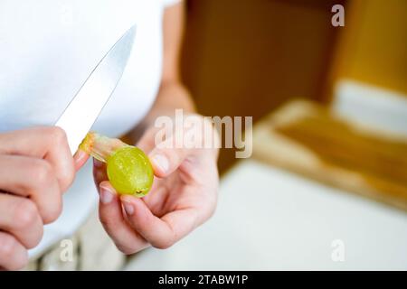 Vue rapprochée de dessus des mains blanches pelant et enlevant la pelure d'un seul raisin avec un couteau de cuisine Banque D'Images
