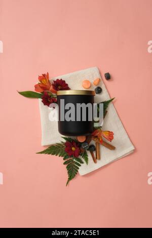 Un pot de bougies parfumées noires non étiquetées est placé à côté de fleurs fraîches, de feuilles vertes et de cannelle sur un fond rose avec un chiffon blanc. Maquette FO Banque D'Images