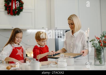 Jeune mère heureuse avec des enfants faisant des biscuits de pain d'épice de Noël et les mettant sur un plateau dans la cuisine. Banque D'Images