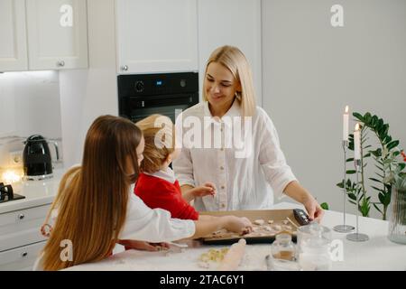 Jeune mère heureuse avec des enfants faisant des biscuits de pain d'épice de Noël et les mettant sur un plateau dans la cuisine. Banque D'Images