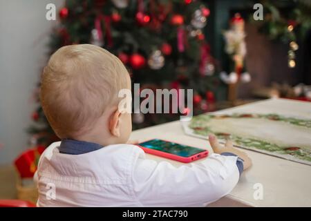 Un petit garçon est assis dans la cuisine décorée pour Noël à la table et regarde son téléphone portable Banque D'Images