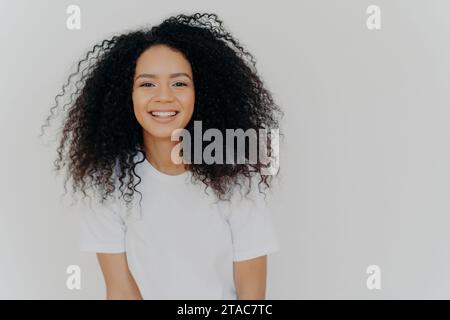 Femme noire radieuse avec des cheveux bouclés volumineux dans un tee-shirt blanc souriant chaleureusement. Banque D'Images