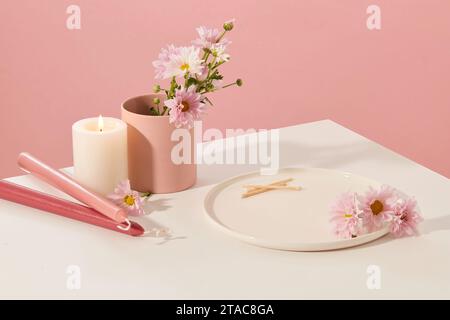 Sur la table blanche, il y a des bougies, des fleurs fraîches, une tasse en céramique et une assiette en céramique avec un fond rose. Espace pour la présentation du produit. Thème beauté Banque D'Images