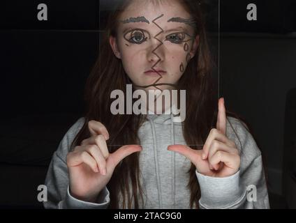 Adolescente tient une feuille transparent de perspex avec le dessin d'un demi-visage sur son propre visage pour montrer différentes émotions et sentiments Banque D'Images