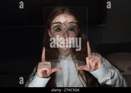 Adolescente tient une feuille transparent de perspex avec le dessin d'un demi-visage sur son propre visage pour montrer différentes émotions et sentiments Banque D'Images